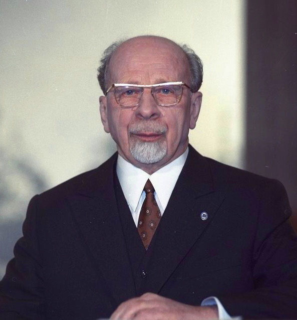 Retrato de Walter Ulbricht, líder da Alemanha Oriental. Ele é um homem branco, calvo, tem cavanhaque grisalho e usa óculos. Ele está vestido com terno e gravata e olha para a frente.