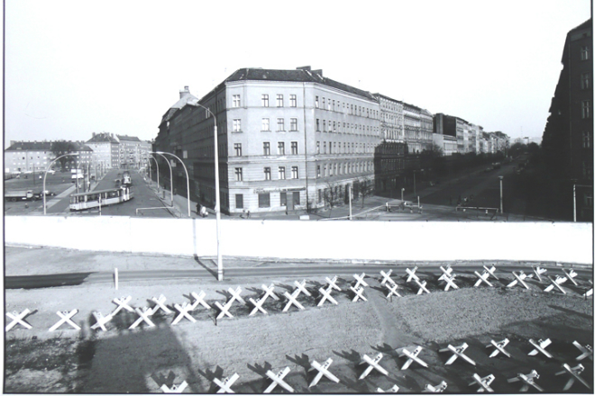 Foto da rua Bernau, em Berlim, dividida ao meio pelo Muro de Berlim. É possível ver um muro branco erguido e cavaletes para impedir a passagem de pessoas. Ao fundo, um bonde dobra a esquina.
