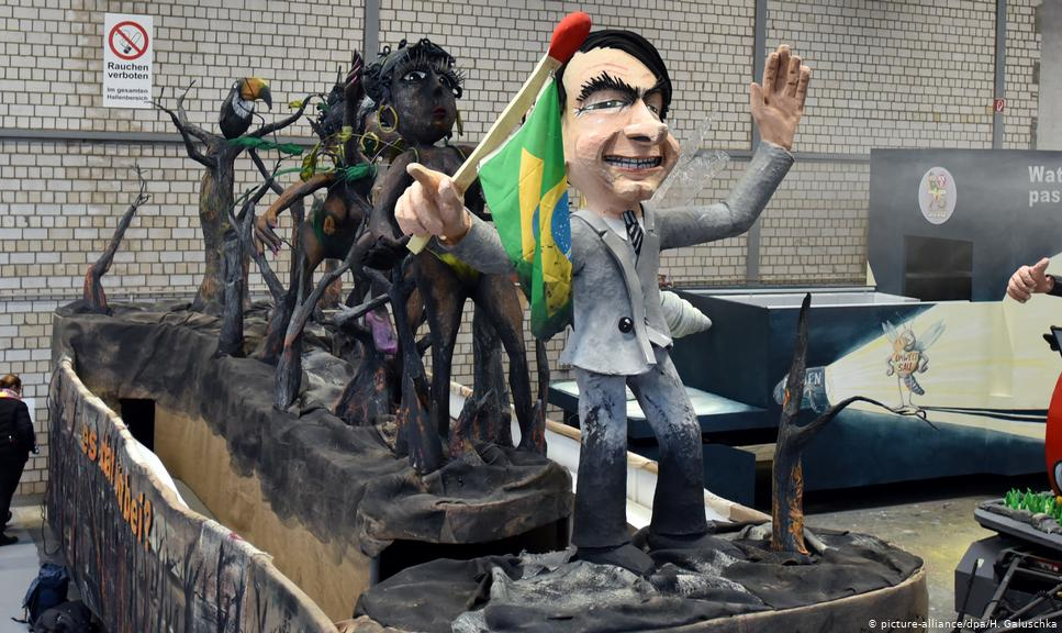 No Carnaval de Colônia, boneco do presidente porta bandeira presa a palito de fósforo diante de passistas carbonizadas